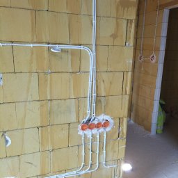 Daniel Lisiecki - Instalacja elektryczna w trakcie montażu na ścianie z bloczków, widoczne przewody w białych peszlach, puszki instalacyjne w kolorze pomarańczowym oraz fragment ubrania roboczego w tle.