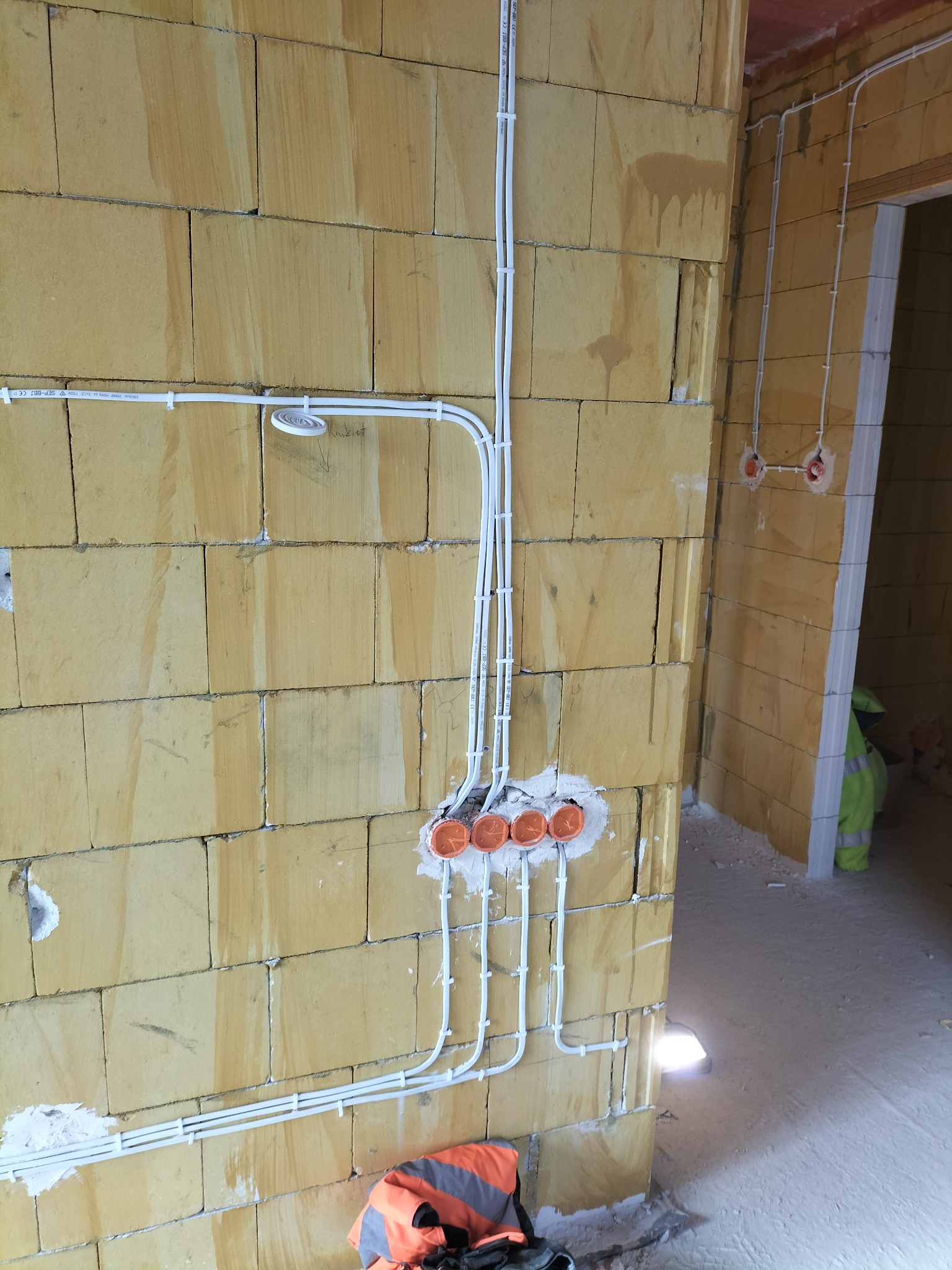 Instalacja elektryczna w trakcie montażu na ścianie z bloczków, widoczne przewody w białych peszlach, puszki instalacyjne w kolorze pomarańczowym oraz fragment ubrania roboczego w tle.