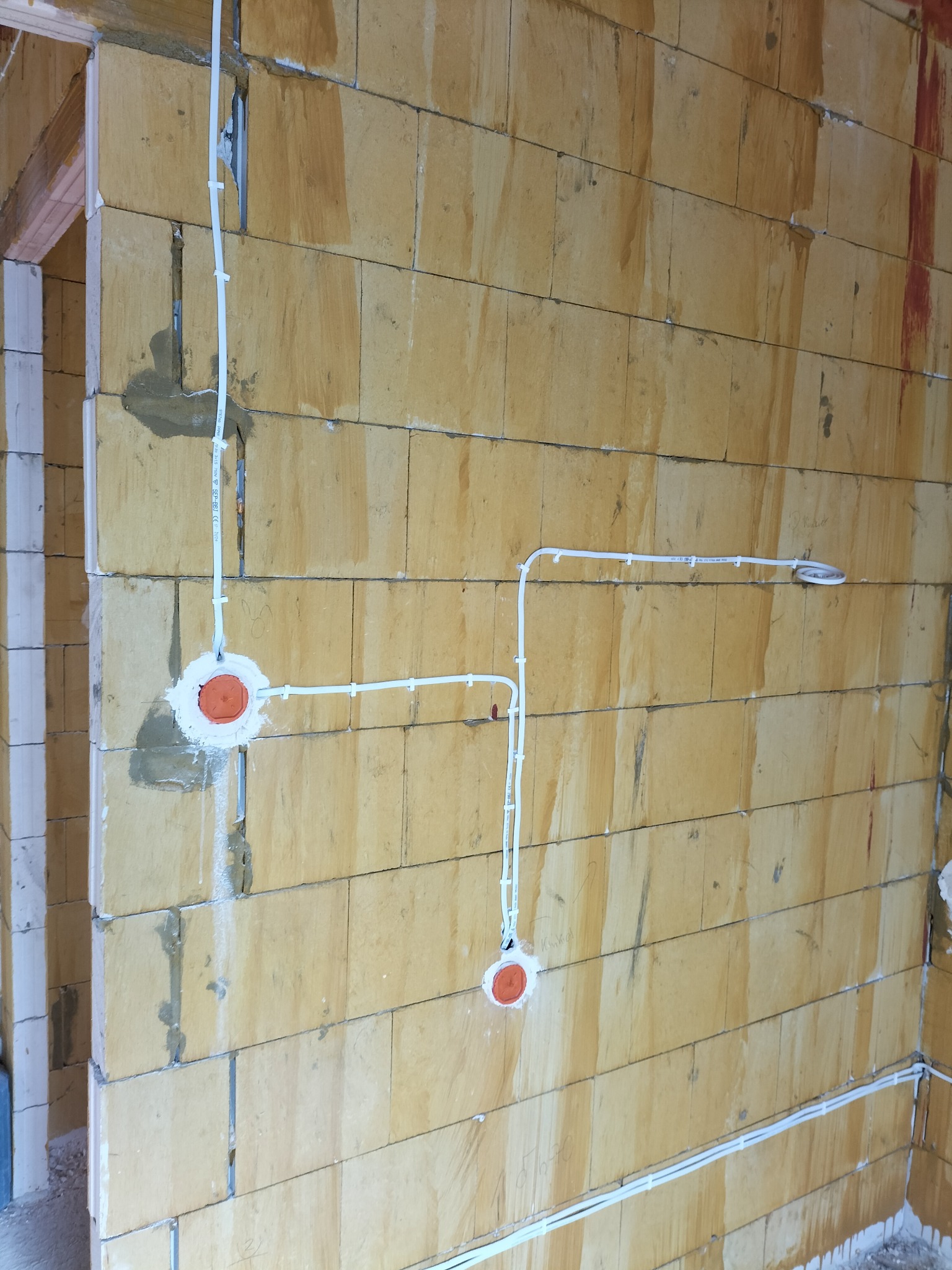 Wewnętrzna ściana z pustaków, z widoczną instalacją elektryczną: białe przewody prowadzone po powierzchni, puszki instalacyjne z pomarańczowymi dekielkami, mocowane gipsem.