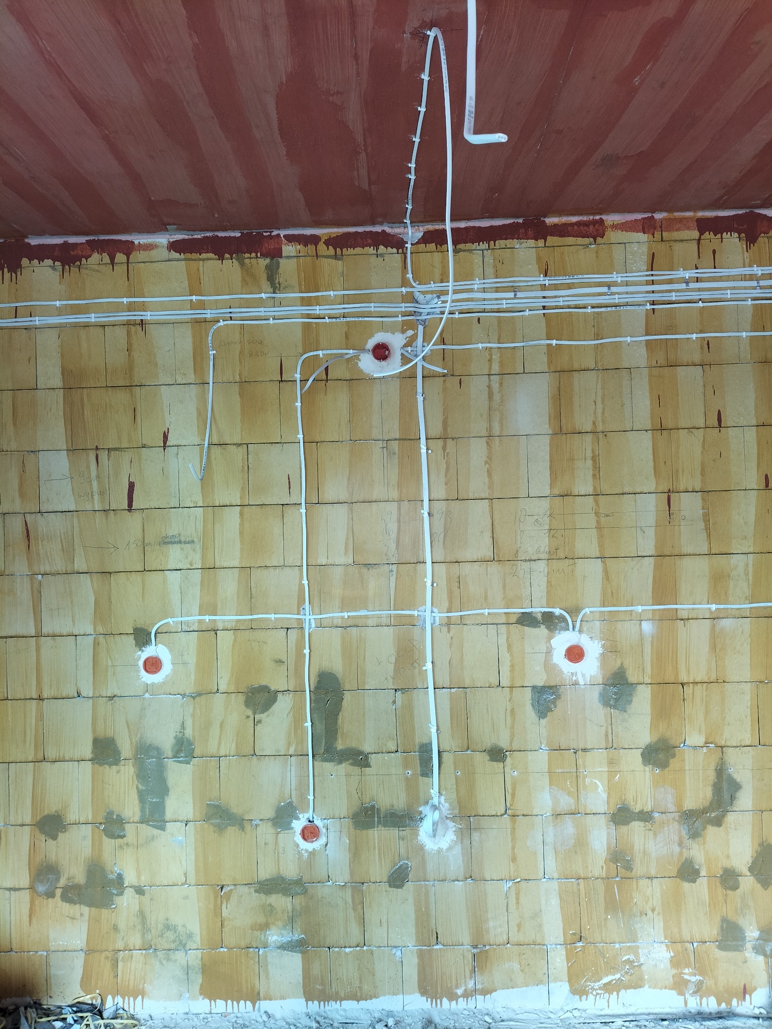 Wewnętrzna instalacja elektryczna: przewody w białych rurkach mocowane do ściany z pustaków, widoczne puszki instalacyjne z pomarańczowymi dekielkami, ślady zaprawy na ścianie.