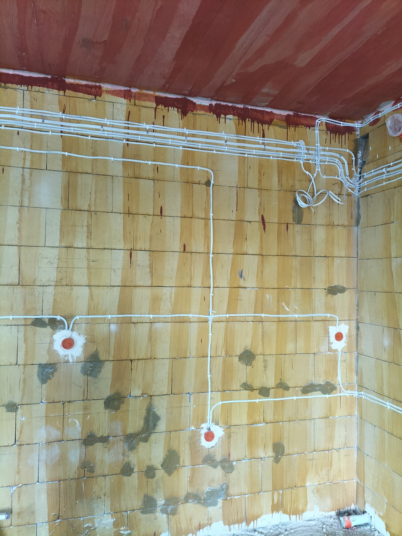 Surowy mur z widocznymi przewodami elektrycznymi w białych rurkach, puszki instalacyjne zamontowane w ścianie, sufit w kolorze ceglastym z zaciekami farby.