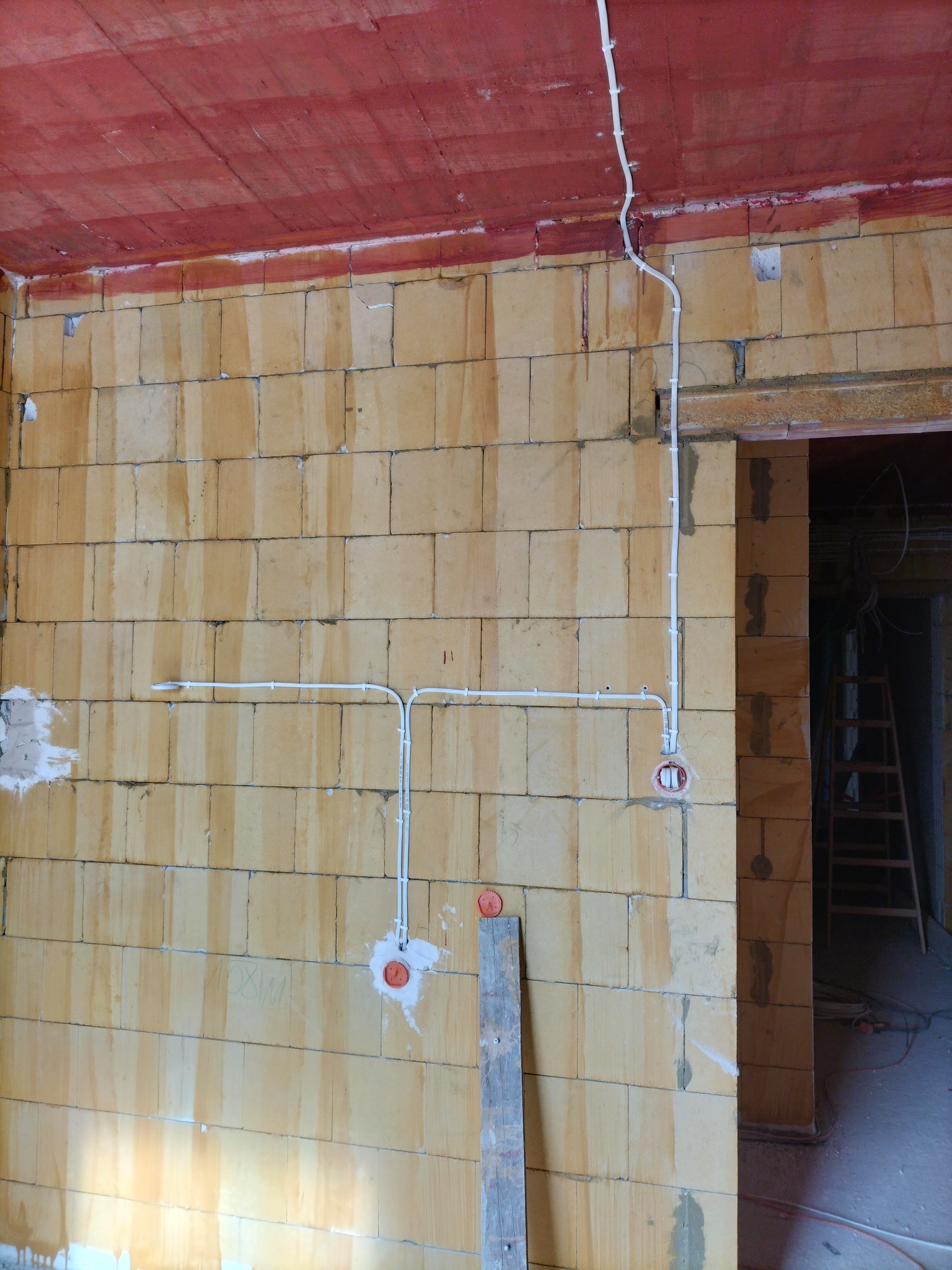 Wewnętrzna instalacja elektryczna w surowym budynku z ceglanymi ścianami i czerwonym sufitem, widoczne białe przewody i puszki podtynkowe przed tynkowaniem.