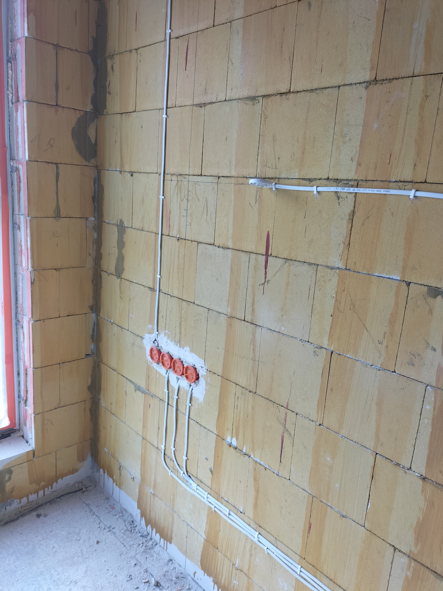 Surowa ściana z betonu komórkowego z zamontowanymi, pomarańczowymi puszkami elektrycznymi i białymi przewodami w trakcie instalacji.