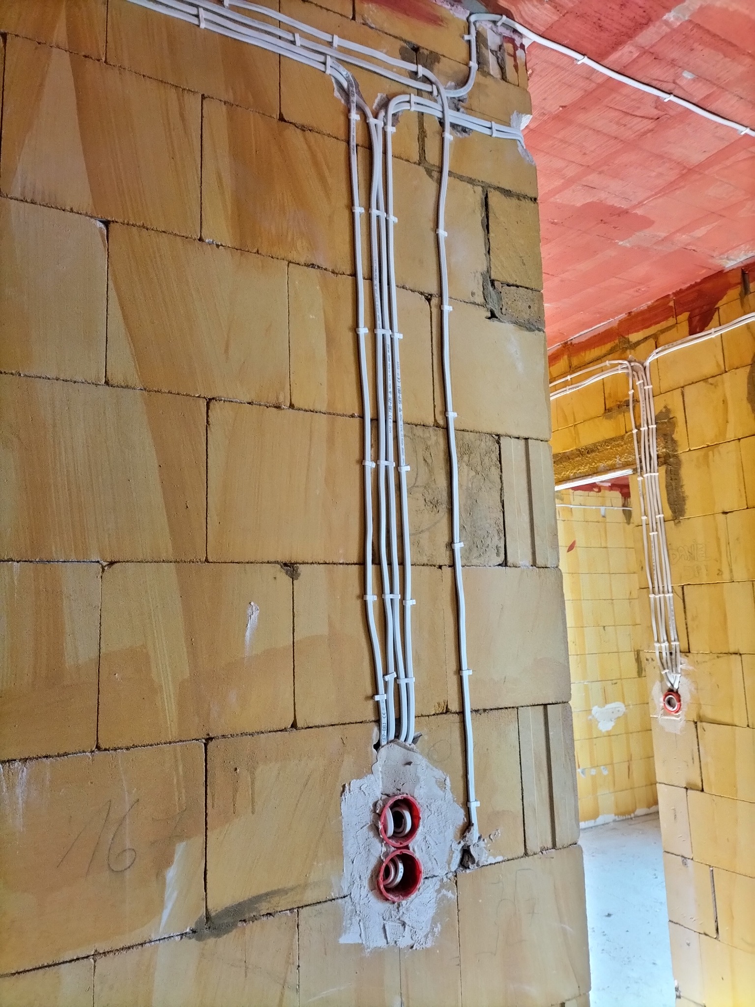 Białe kable elektryczne prowadzone po ścianie z bloczków, zabezpieczone uchwytami, z widocznymi czerwonymi puszkami instalacyjnymi w szarym tynku.