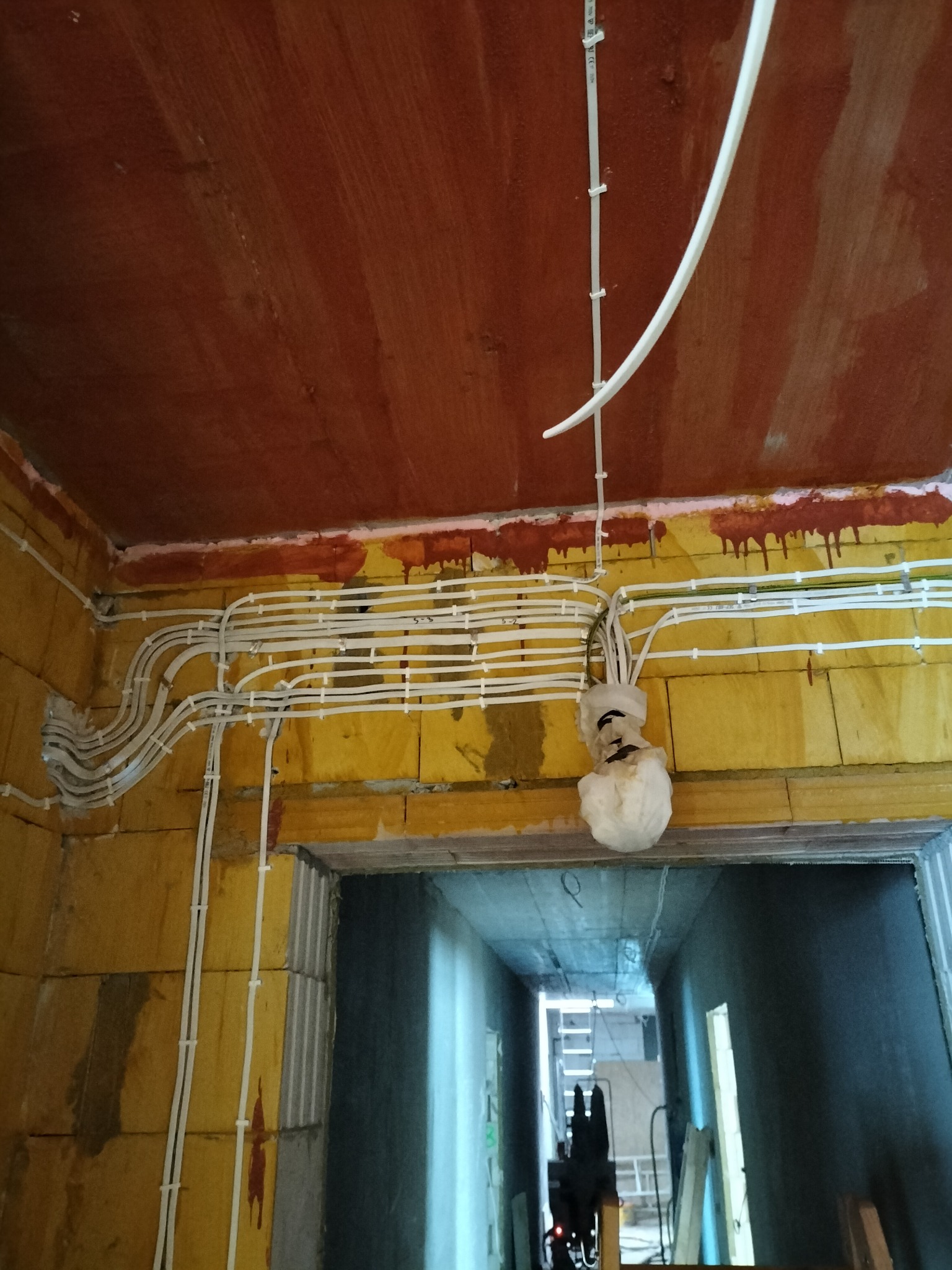 Rozprowadzone przewody elektryczne w białych rurkach PCV, mocowane klamrami do surowych ścian z pustaków, widok na korytarz w trakcie remontu, zabezpieczone kable zwisające z sufitu.
