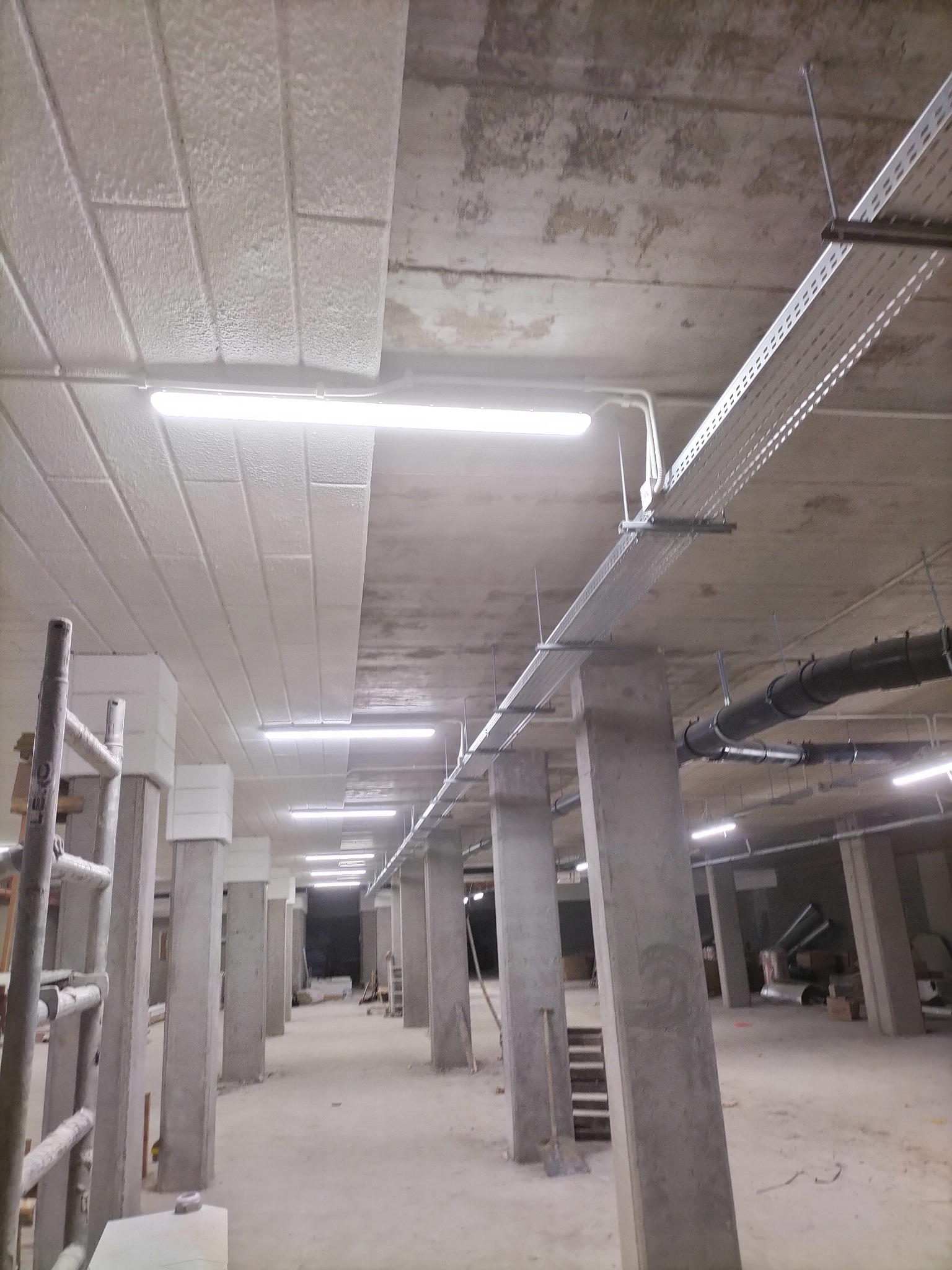 Wnętrze w stanie surowym z widoczną instalacją oświetleniową, korytkami kablowymi pod sufitem i betonowymi kolumnami.