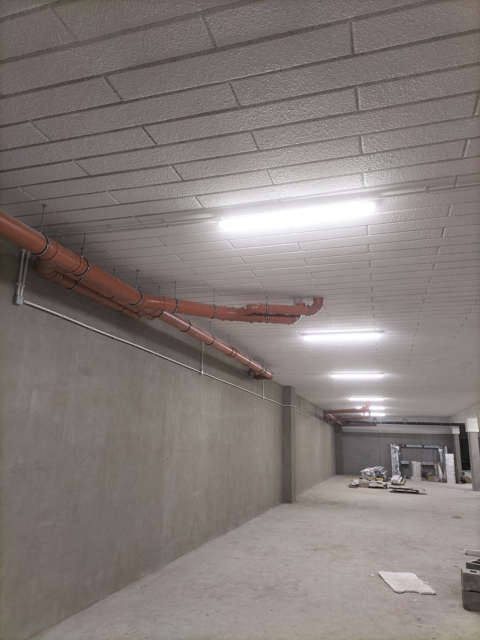 Pomieszczenie w stanie surowym z widocznymi pomarańczowymi rurami wentylacyjnymi podwieszonymi do sufitu oraz oświetleniem ledowym.