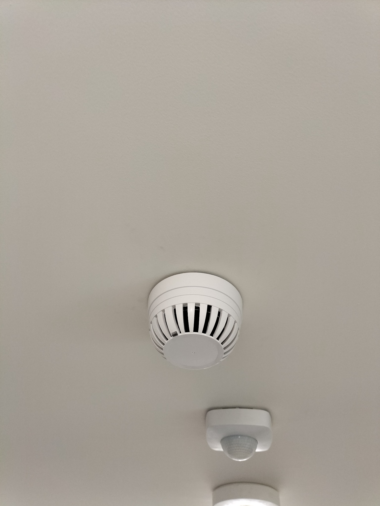 Biały czujnik dymu z kratką wentylacyjną i detektorem ruchu zamontowany na suficie. Widok od dołu.