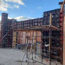 Szare szalunki fundamentowe z czerwonymi elementami mocującymi, częściowo wypełnione cegłą, widoczne rusztowanie i drabina na placu budowy w słoneczny dzień.