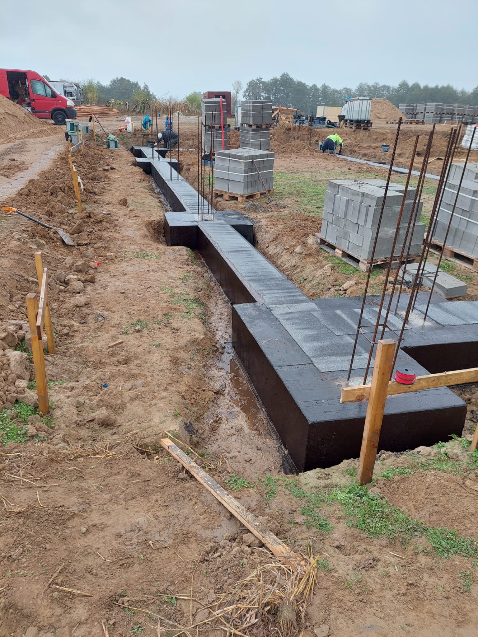 Ciemna, zabezpieczona przed wilgocią, izolacja fundamentowa w wykopie, wystające pręty zbrojeniowe, palety z bloczkami betonowymi w tle, ziemia wokół wykopu.