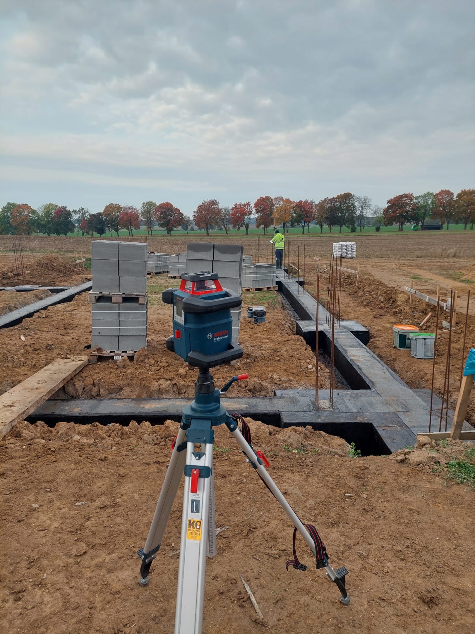 Niwelator laserowy Bosch na statywie, widoczne fundamenty zbrojone i zabezpieczone izolacją, palety z bloczkami betonowymi, pracownik w odblaskowej kamizelce w tle.