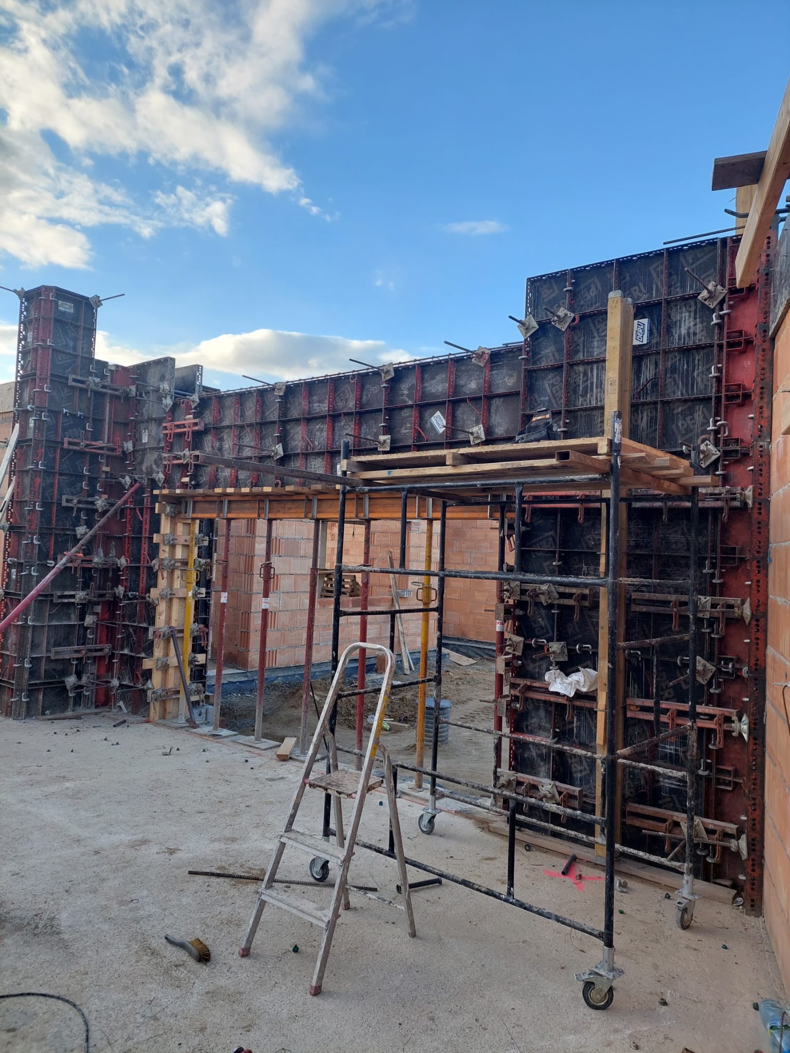 Szare szalunki fundamentowe z czerwonymi elementami mocującymi, częściowo wypełnione cegłą, widoczne rusztowanie i drabina na placu budowy w słoneczny dzień.