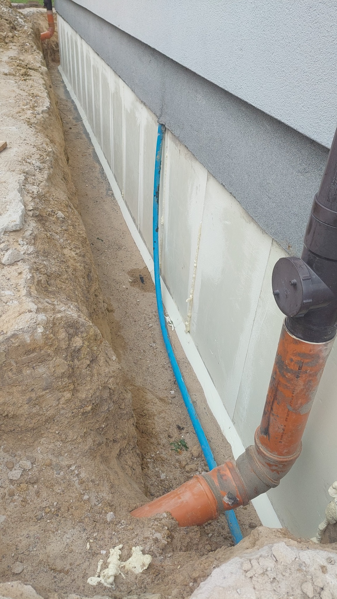 Wykop wzdłuż fundamentu budynku z widoczną izolacją termiczną i wystającą rurą kanalizacyjną z pomarańczowego tworzywa, oraz niebieski przewód w ziemi.