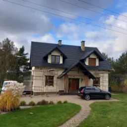 Dom jednorodzinny z nowym, ciemnym dachem, obudowany rusztowaniami, z samochodem zaparkowanym na podjeździe z kostki brukowej.