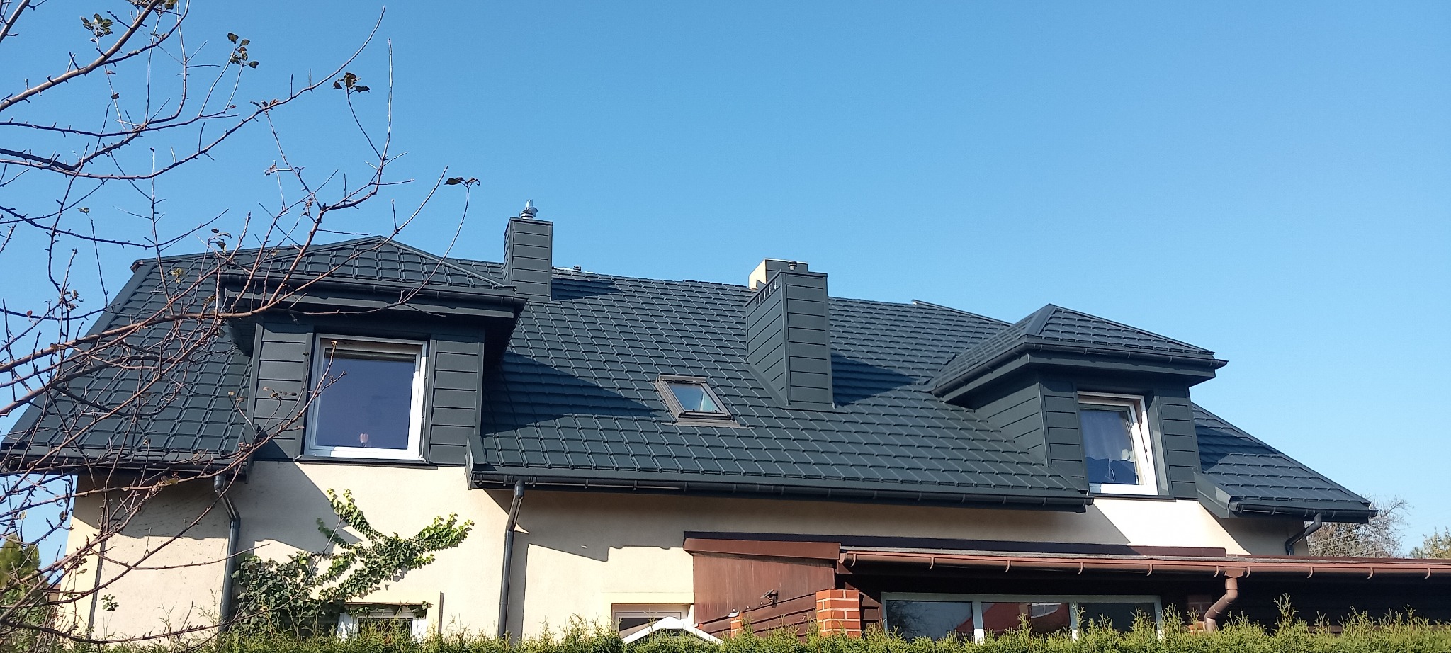 Dom z nowym, ciemnoszarym dachem z dachówki ceramicznej, widoczne lukarny, kominy i rynny, na tle błękitnego nieba.