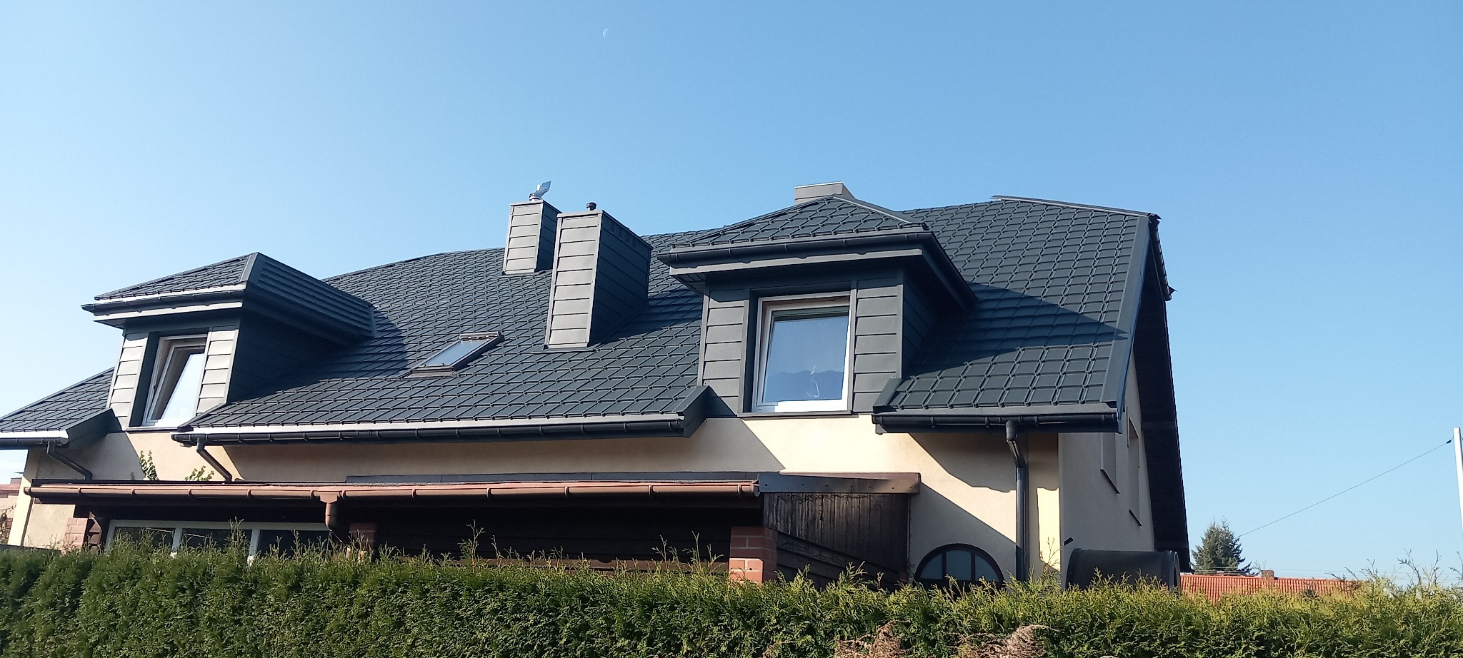 Dom jednorodzinny z ciemnoszarą dachówką, rynnami i obróbkami blacharskimi, widoczne kominy i okna dachowe, na dachu antena w kształcie wiatrowskazu.