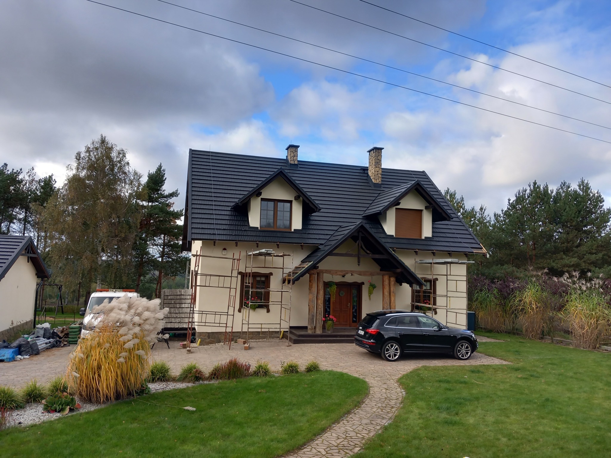 Dom jednorodzinny z nowym, ciemnym dachem, obudowany rusztowaniami, z samochodem zaparkowanym na podjeździe z kostki brukowej.