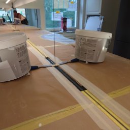 MAR-BUD CONSTRUCTION - Przygotowanie powierzchni: Pomalowana taśma maskująca wyznacza linie na zabezpieczonej kartonem podłodze, widoczne dwa wiadra z farbą i szpachelki, w tle okno z powieszonymi notatkami.