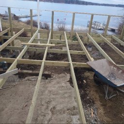 MAR-BUD CONSTRUCTION - Konstrukcja drewnianego tarasu w trakcie budowy, widok na jezioro, taczka z kamieniami i poziomica. Elementy konstrukcji są jasnego drewna.