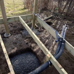 MAR-BUD CONSTRUCTION - Konstrukcja drewniana w trakcie budowy na ziemi z widocznymi instalacjami, podsypką z kamienia i elementami mocującymi.