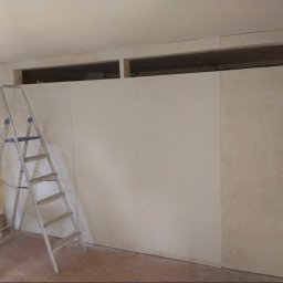 MAR-BUD CONSTRUCTION - Wnętrze w trakcie prac remontowych: ściana z płyt gipsowo-kartonowych z otworami, drabina aluminiowa oparta o ścianę, fragment ściany z resztkami starej farby.