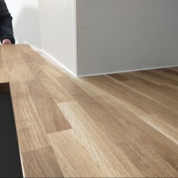 MAR-BUD CONSTRUCTION - Montaż blatu kuchennego z drewnopodobnego laminatu, widoczne łączenie z białą zabudową, osoba w kurtce w tle.