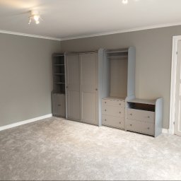 MAR-BUD CONSTRUCTION - Pusty pokój z szarą wykładziną i szarymi meblami modułowymi: regał z półkami, szafa przesuwna i komoda z szufladami, białe drzwi.