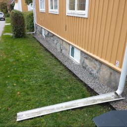 MAR-BUD CONSTRUCTION - Żółta elewacja domu z kamienną podmurówką, obok trawnik z rynną odprowadzającą wodę.