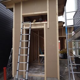 MAR-BUD CONSTRUCTION - Wznoszenie szkieletu małego budynku gospodarczego z widoczną konstrukcją ścian, dachu i drabiny opartą o ścianę. Wewnątrz widoczny częściowo szkielet konstrukcji.