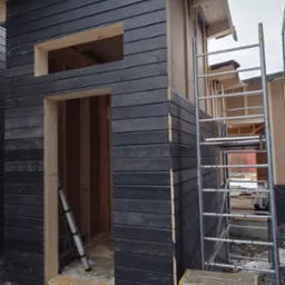 Surowa elewacja z ciemnego drewna w trakcie budowy, widoczne ramy okienne i drzwiowe, rusztowanie aluminiowe i drabina, teren budowy z resztkami materiałów.