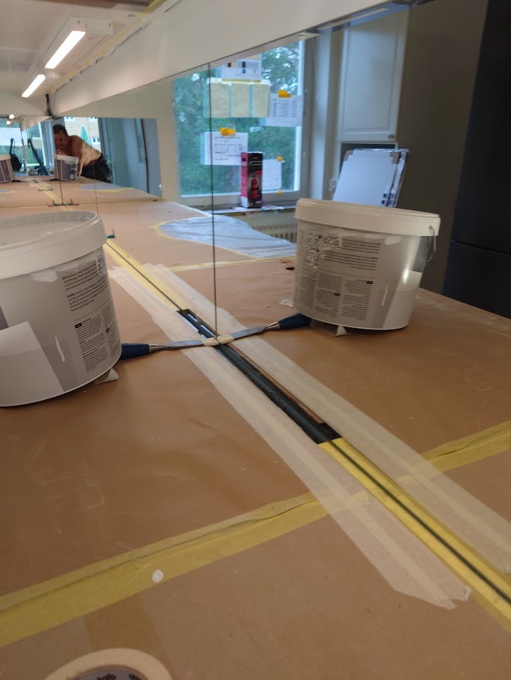 Przygotowanie powierzchni: Pomalowana taśma maskująca wyznacza linie na zabezpieczonej kartonem podłodze, widoczne dwa wiadra z farbą i szpachelki, w tle okno z powieszonymi notatkami.