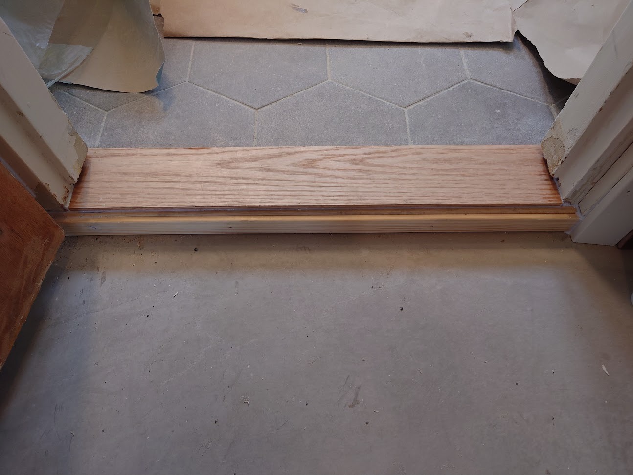 Nowo zamontowany drewniany próg drzwiowy z widocznymi elementami podłogi wyłożonej szarymi, sześciokątnymi płytkami oraz fragmentem surowego betonu, zabezpieczony papierem malarskim przy framudze.