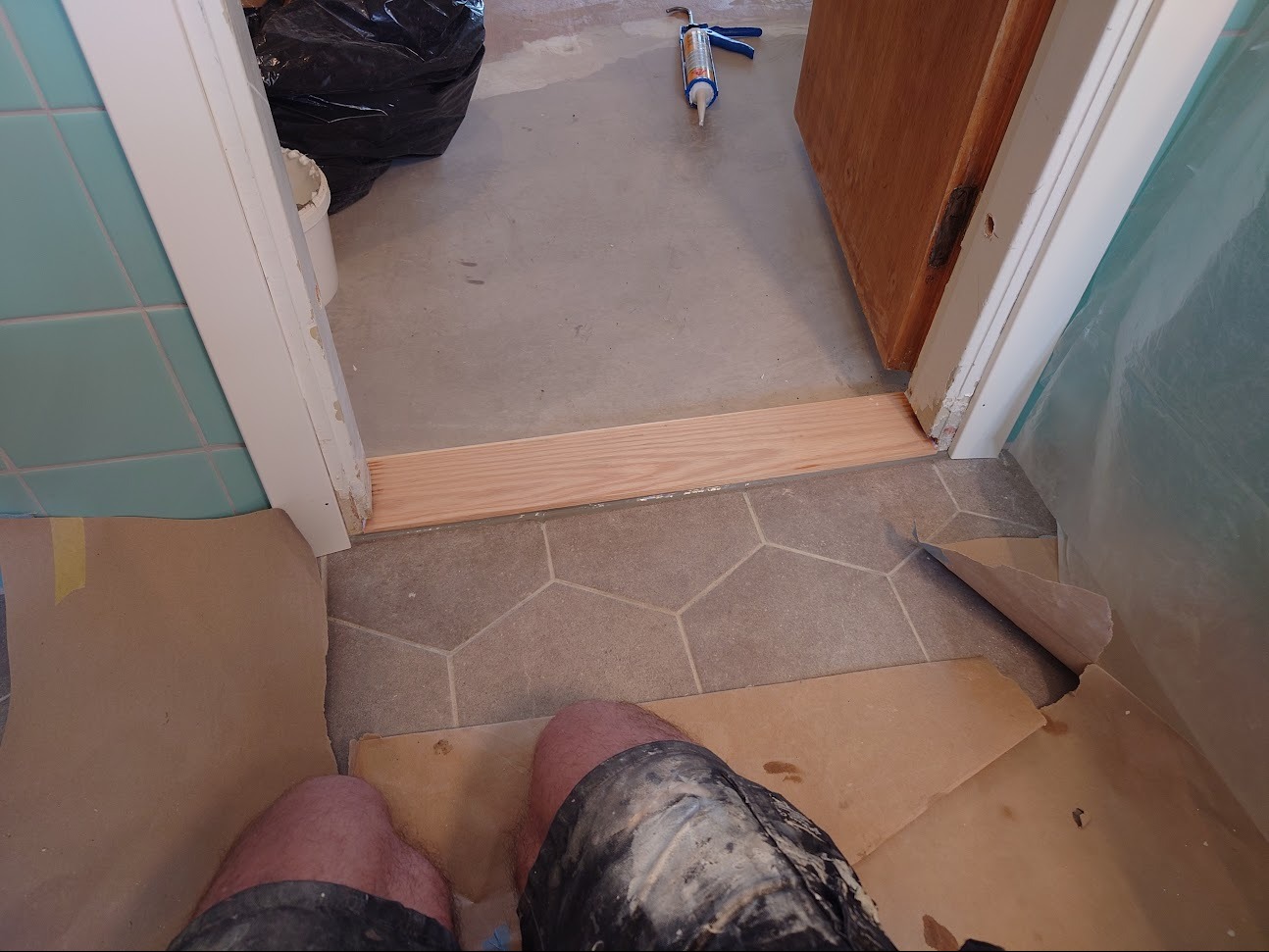 Montaż drewnianego progu w odnawianej łazience z turkusowymi płytkami, widoczny aplikator do silikonu i zabezpieczenia podłogi.