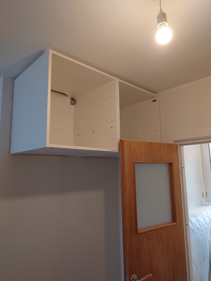Montaż białych szafek kuchennych pod sufitem w Bydgoszczy, widoczna drewniana ościeżnica drzwi z szybą i surowa żarówka