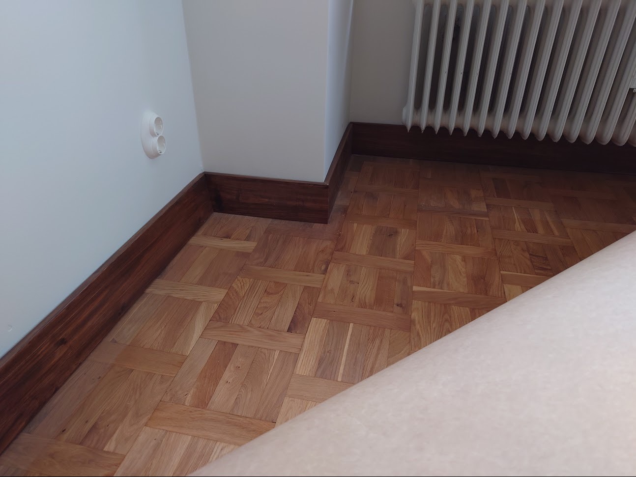 Parkiet ułożony w jodełkę z ciemną listwą przypodłogową, widoczny fragment grzejnika i gniazdko elektryczne.