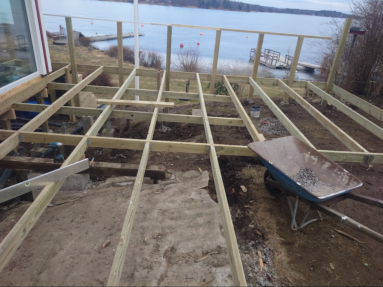 Konstrukcja drewnianego tarasu w trakcie budowy, widok na jezioro, taczka z kamieniami i poziomica. Elementy konstrukcji są jasnego drewna.