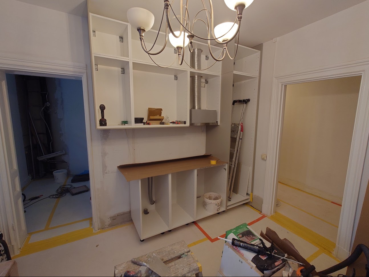 Wnętrze kuchni w trakcie montażu zabudowy meblowej, widoczne białe szafki bez frontów, okap, drabina oparta o ścianę, zabezpieczona podłoga taśmą malarską, narzędzia i materiały budowlane.