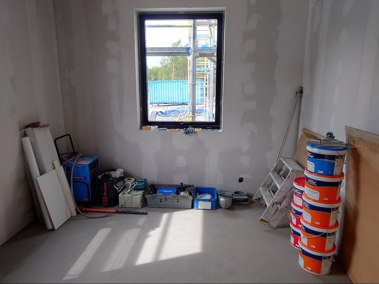 Wnętrze w trakcie prac wykończeniowych: ściany po nałożeniu gładzi, okno z widokiem na rusztowanie i kontener, na podłodze narzędzia i wiaderka z farbą.