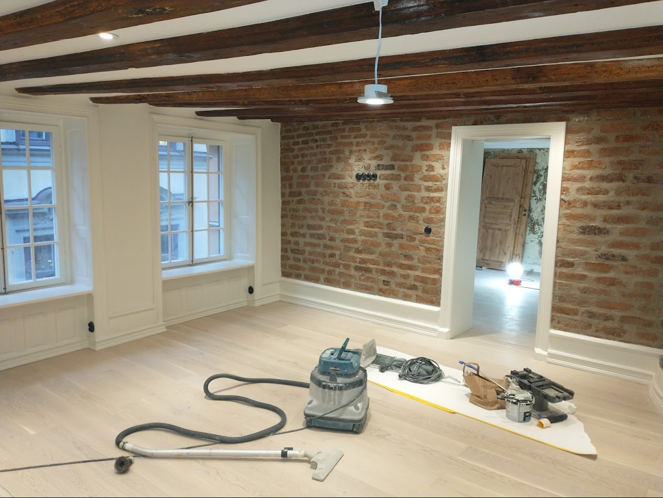 Jasne panele podłogowe po cyklinowaniu w pokoju z ceglaną ścianą i drewnianym sufitem, widoczny odkurzacz przemysłowy i narzędzia na folii ochronnej.