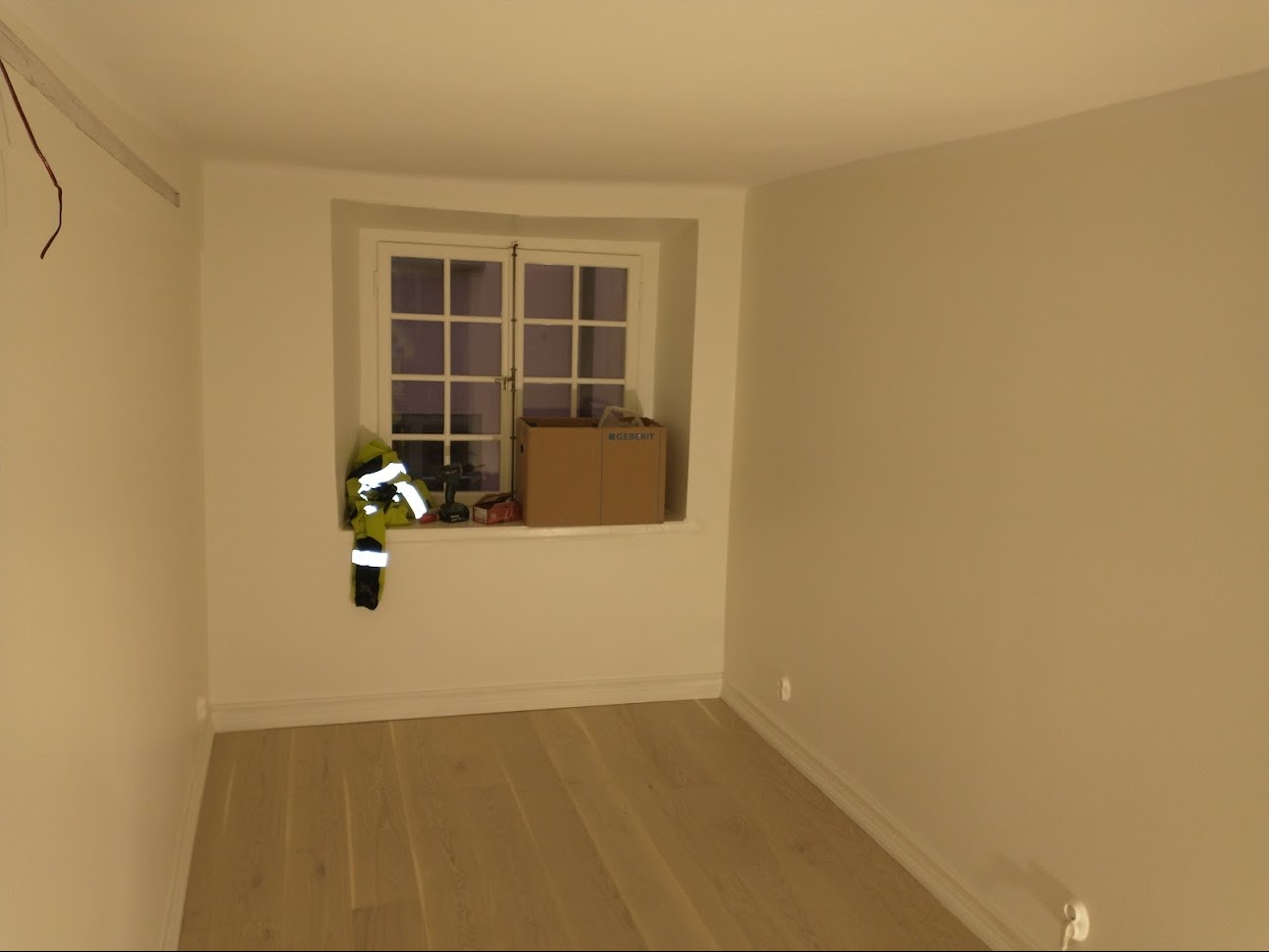 Pusty pokój po remoncie: świeżo położone panele podłogowe, pomalowane na biało ściany, narzędzia i karton na parapecie.