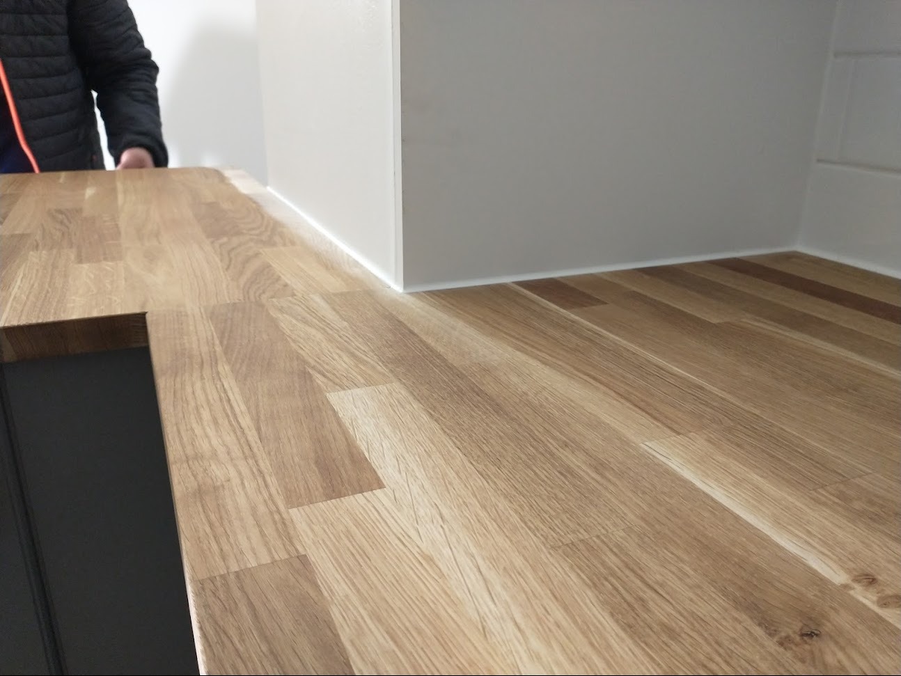 Montaż blatu kuchennego z drewnopodobnego laminatu, widoczne łączenie z białą zabudową, osoba w kurtce w tle.