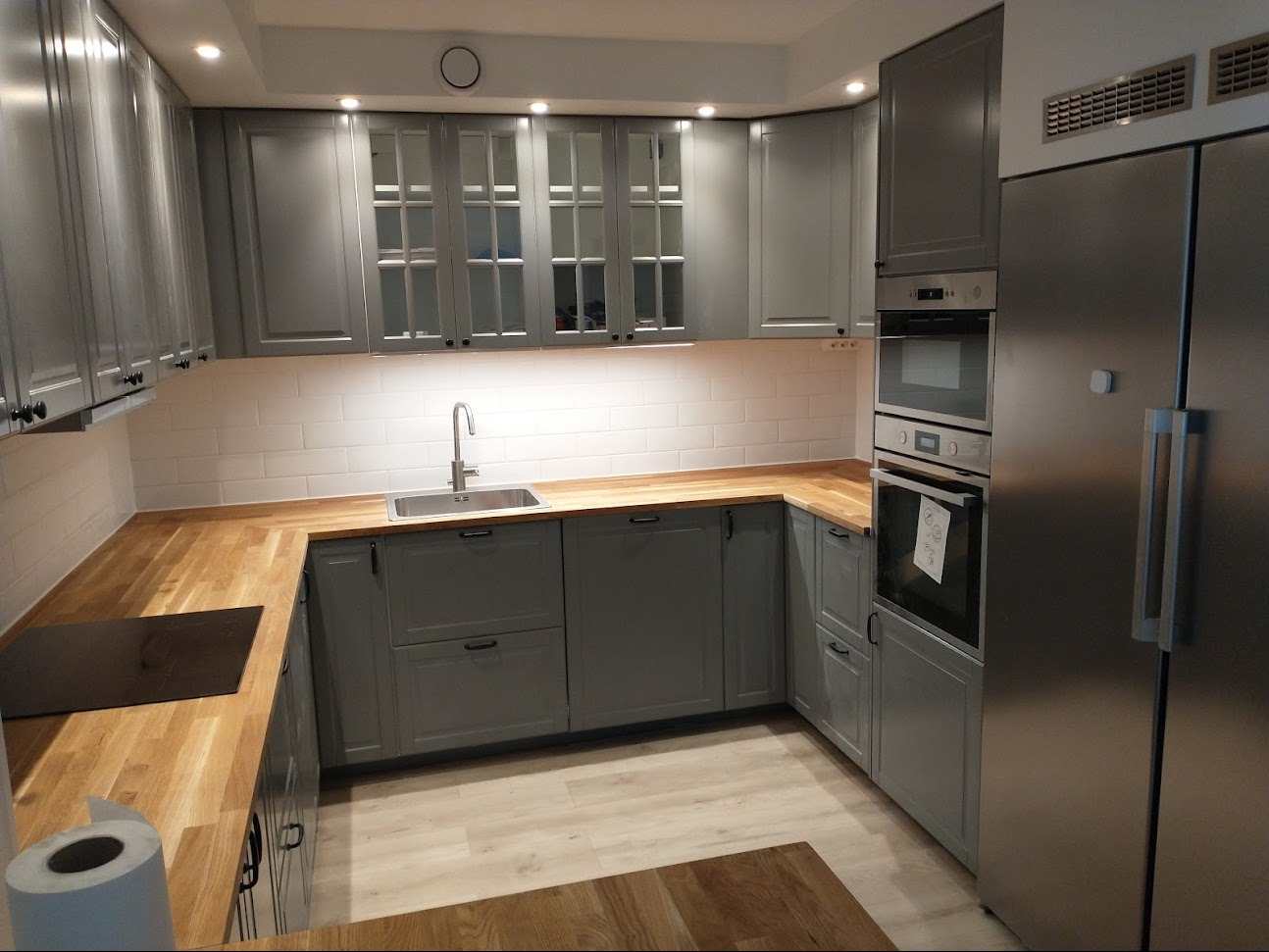 Szara, nowocześnie urządzona kuchnia z drewnianymi blatami, białymi płytkami na ścianie i srebrną lodówką.