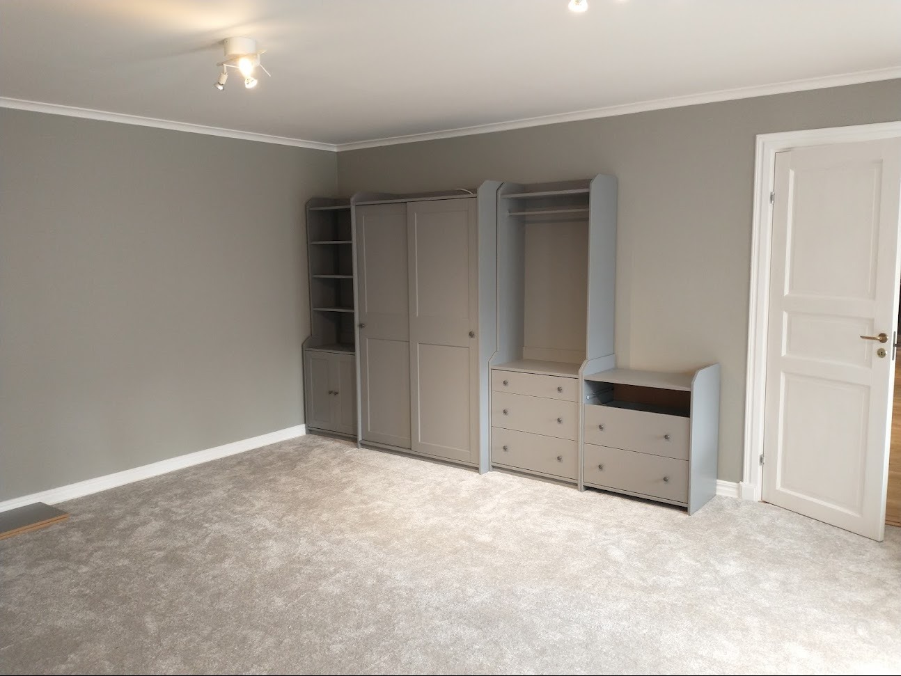 Pusty pokój z szarą wykładziną i szarymi meblami modułowymi: regał z półkami, szafa przesuwna i komoda z szufladami, białe drzwi.
