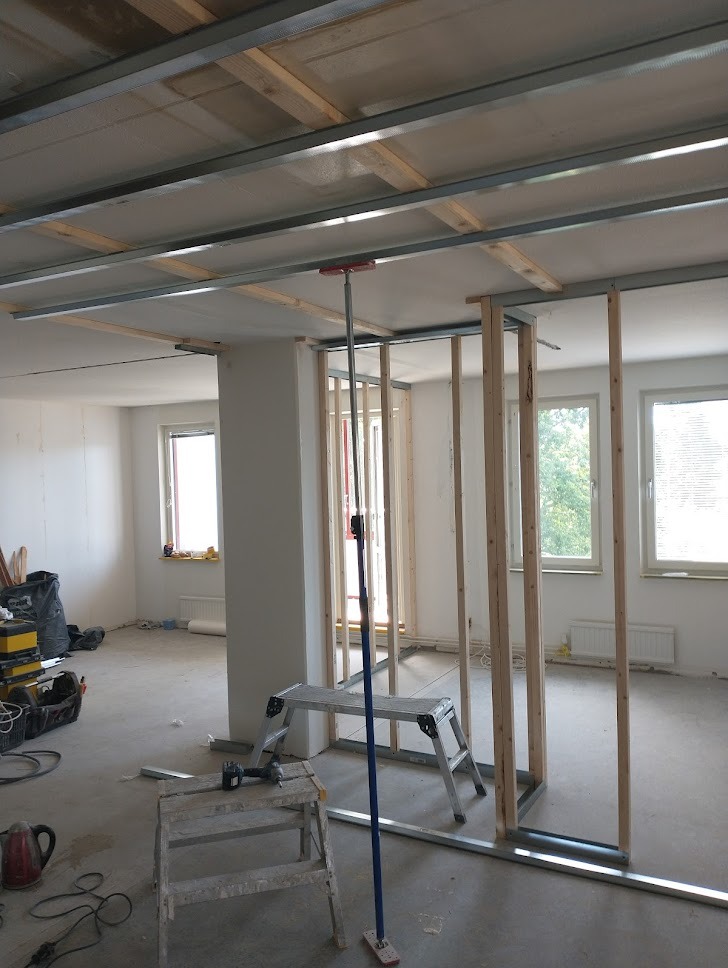 Wnętrze w trakcie remontu: konstrukcja ścian działowych z drewnianych belek i metalowych profili, podwieszany sufit z płyt gipsowych, drabina i narzędzia na podłodze, widok przez okno na zieleń.