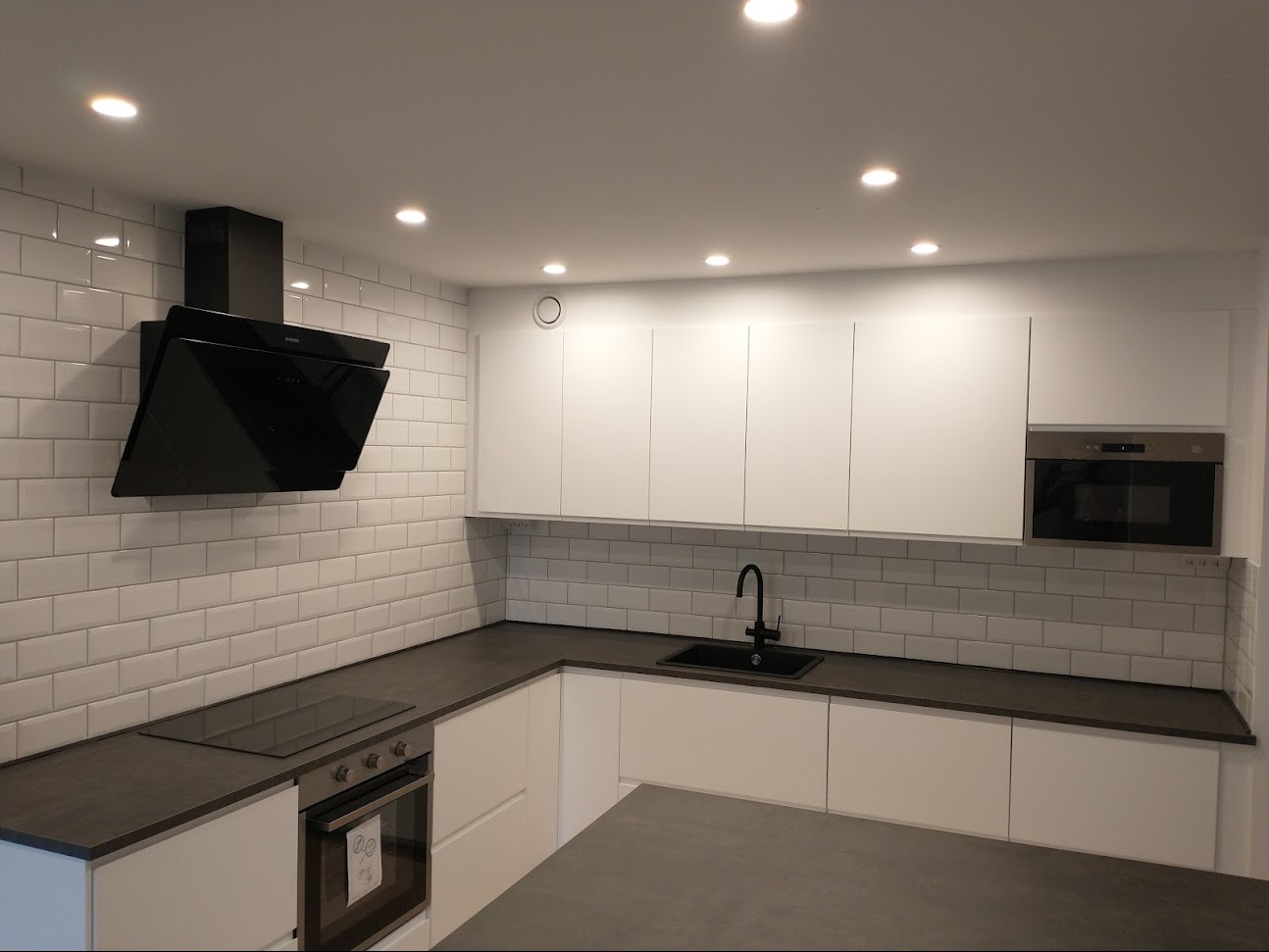 Nowoczesna kuchnia z białymi, minimalistycznymi szafkami, ciemnym blatem i czarną armaturą. Ściana wyłożona białymi, prostokątnymi płytkami imitującymi cegłę. Czarny, skośny okap nad płytą...