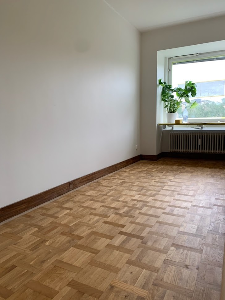 Pusty pokój z parkietem ułożonym we wzór jodełki, białe ściany i grzejnik pod oknem z rośliną doniczkową.