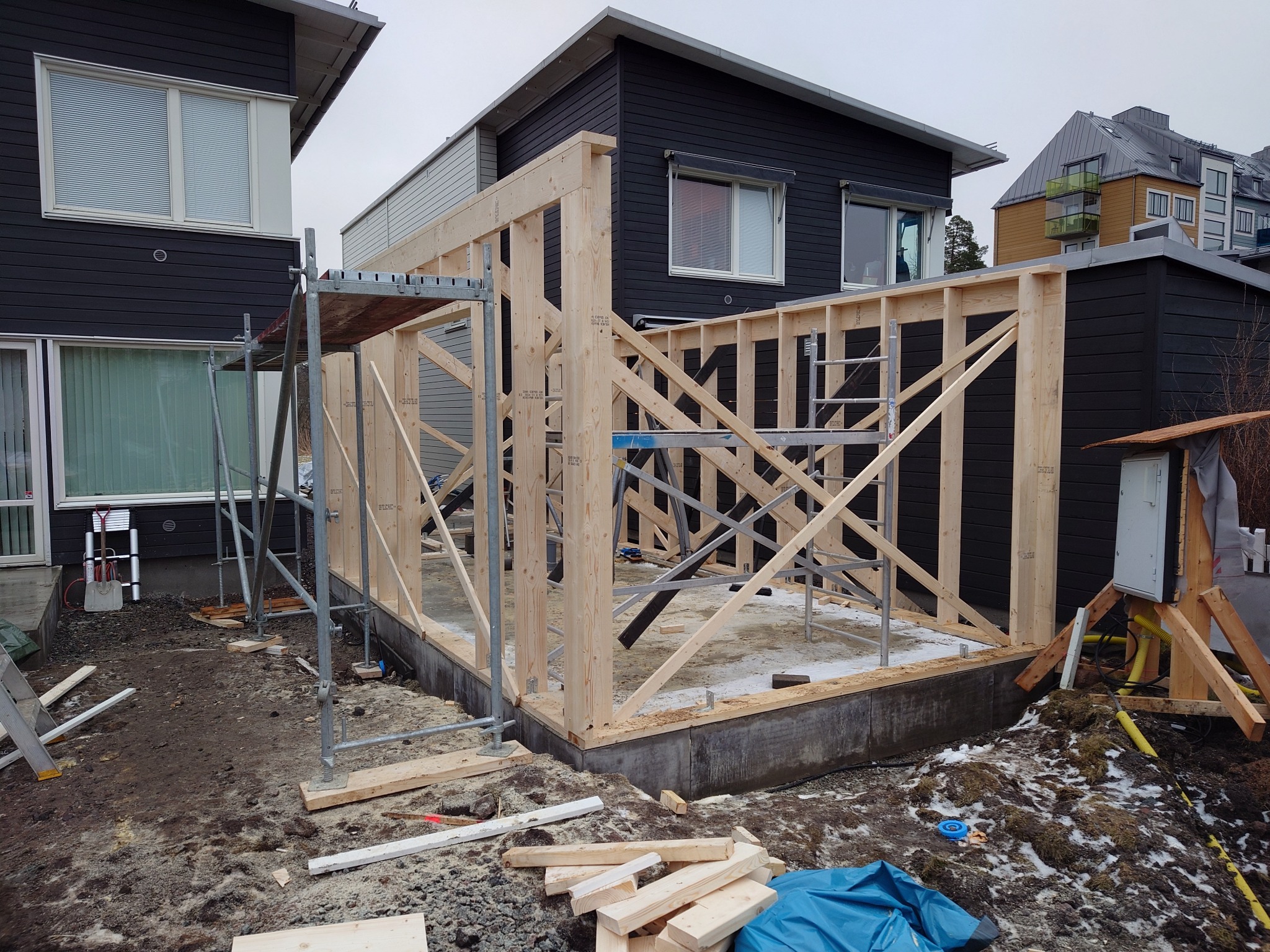 Konstrukcja szkieletowa budynku w trakcie budowy, widoczne rusztowanie, fundament i elementy drewniane, tło stanowią domy z ciemną elewacją.