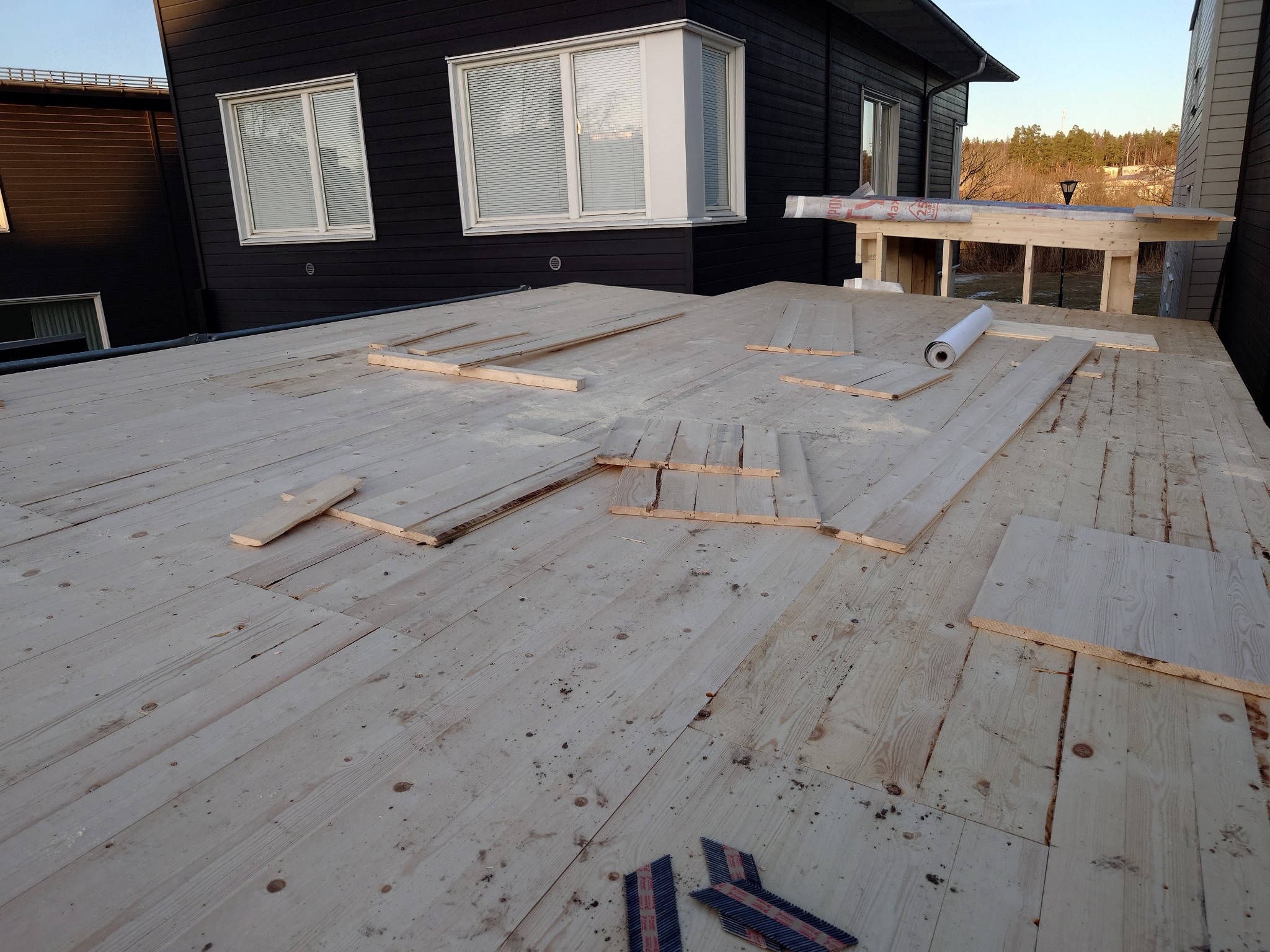 Surowy drewniany taras w trakcie budowy, z widocznymi deskami, rolką izolacji i narzędziami do mocowania, na tle ciemnego domu z oknami.