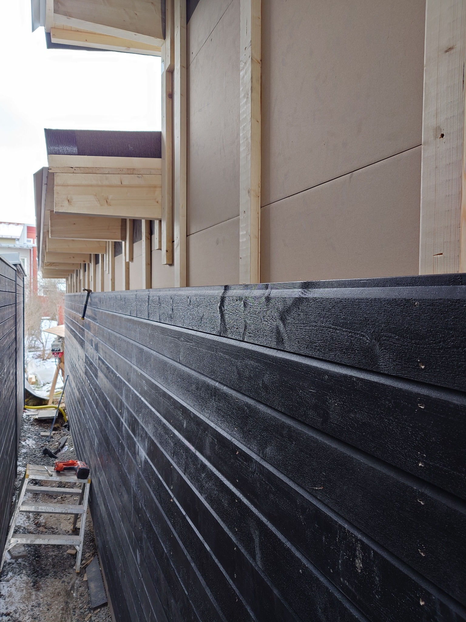 Montaż elewacji z czarnego drewna na budynku w trakcie budowy, widoczna drabina i narzędzia, jasne drewniane elementy konstrukcyjne.