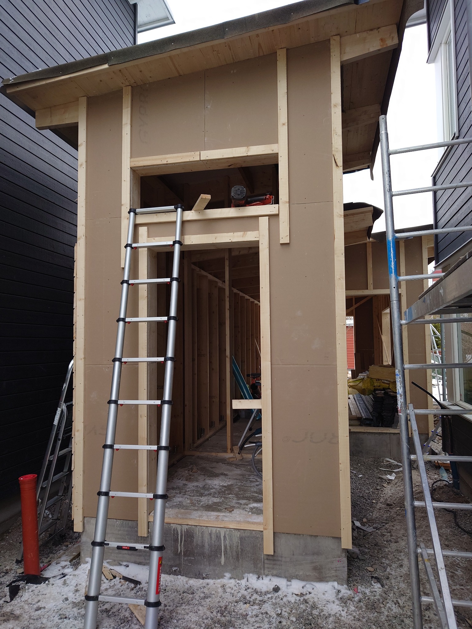Wznoszenie szkieletu małego budynku gospodarczego z widoczną konstrukcją ścian, dachu i drabiny opartą o ścianę. Wewnątrz widoczny częściowo szkielet konstrukcji.